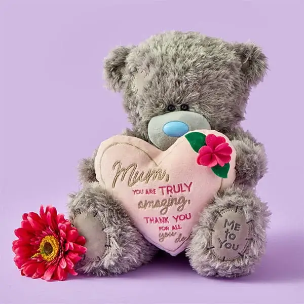 A grey teddy bear holding a heart-shaped cushion with a pink flower decoration, sitting beside a bright pink flower on a pastel purple background.